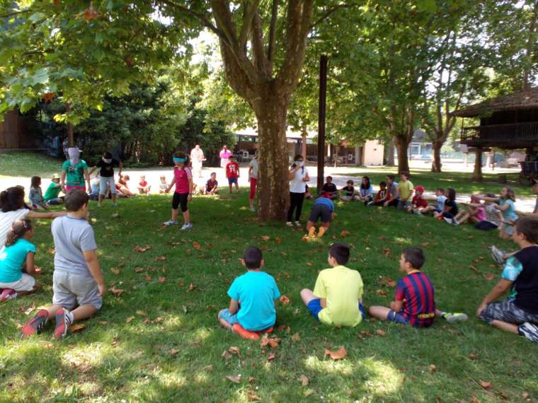 Imagen de niños realizando la actividad de Colonias de Verano en el Muséu Pueblu d`Asturies sentados en círculo en torno a un árbol.