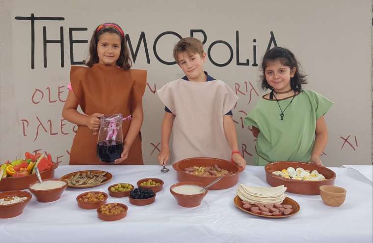 Tres niñas posan en una foto frente a una mesa llena de alimentos en recipientes de barro que, se supone, han cocinado ellas.