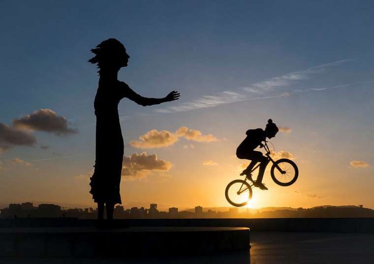 Imagen de una puesta de sol en Gijón en el que se aprecian las siluetas de una ciclista y la estatua de 'La lloca'.