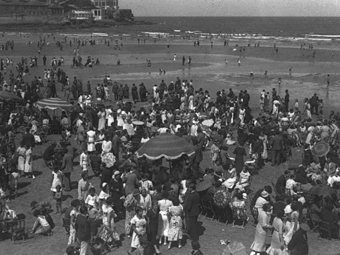 Playa de San Lorenzo con la iglesia de San Pedro y el Cerro de Santa Catalina al fondo el día 28 de agosto de 1934, una jornada dominical en la que el arenal aparece repleto de bañistas. Autor / Taller: Suárez Fernández, Constantino (1899-1983) Archivado: Muséu del Pueblu d'Asturies Colección: Constantino Suárez (FF). 