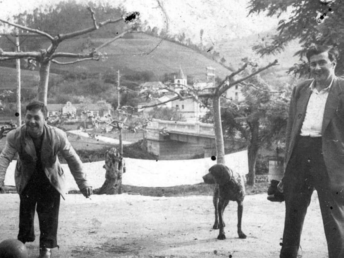 Fotografía dos hombres jugando a la pelota, junto a un perro.